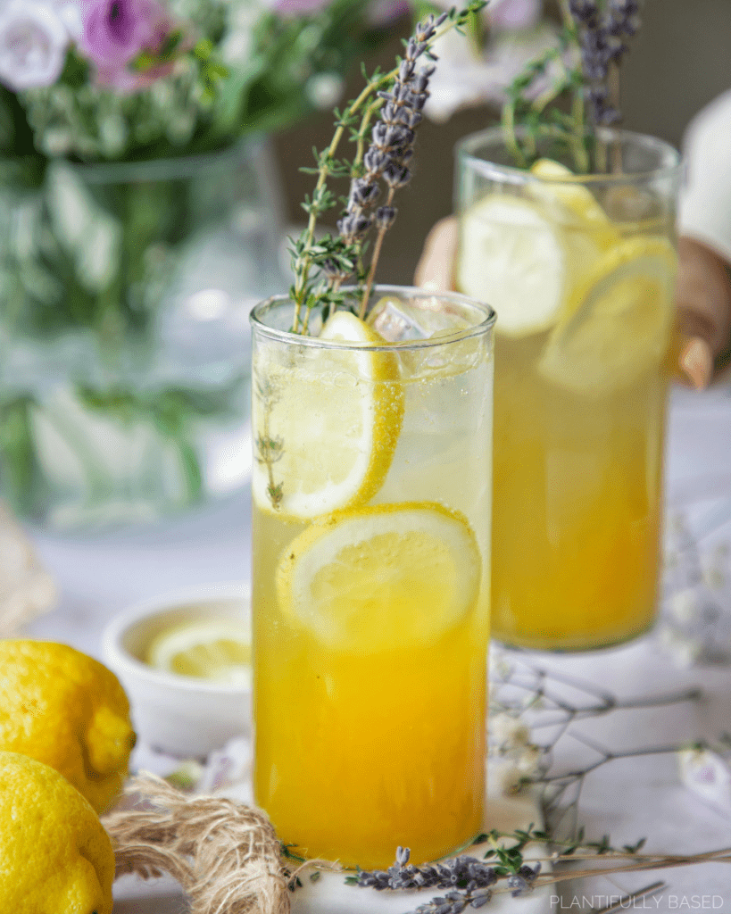 mango passion fruit refresher two glasses one is being held in background. orange yellow drink with lemon slices, fresh thyme and lavender as garnish